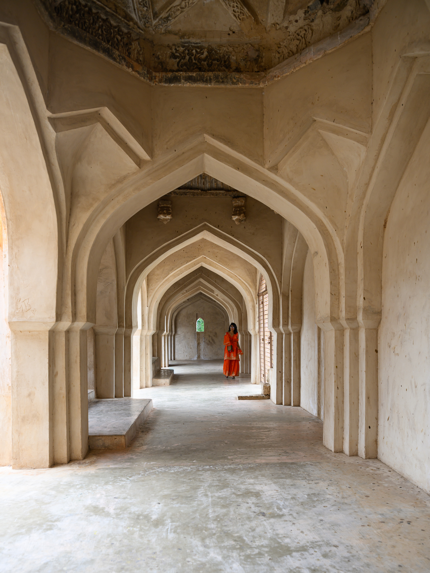 A woman in bright red stands within a long pale corridor of repeating pointed arches.