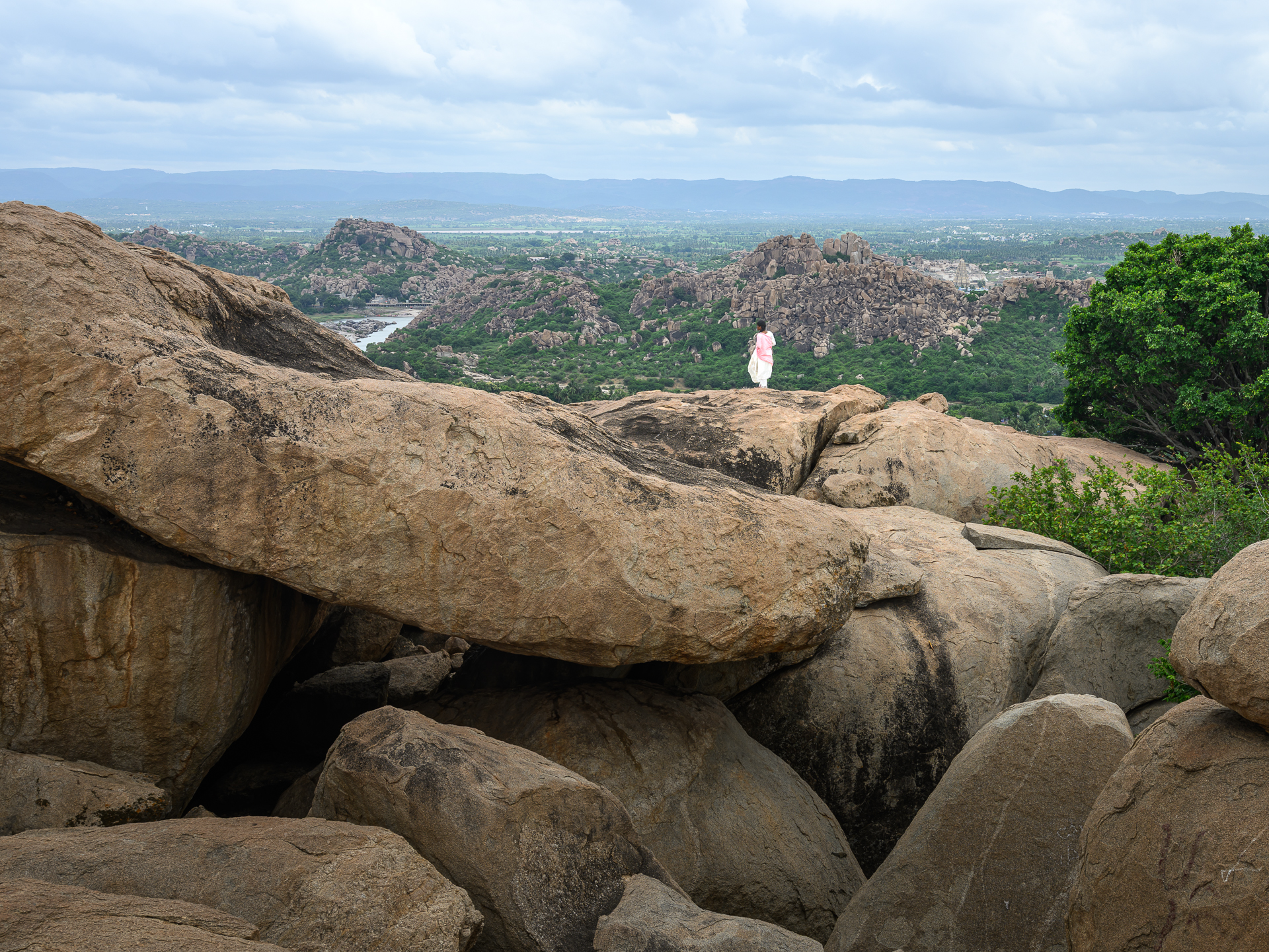 A lone figure in white stands among huge granite boulders above a wide green valley.