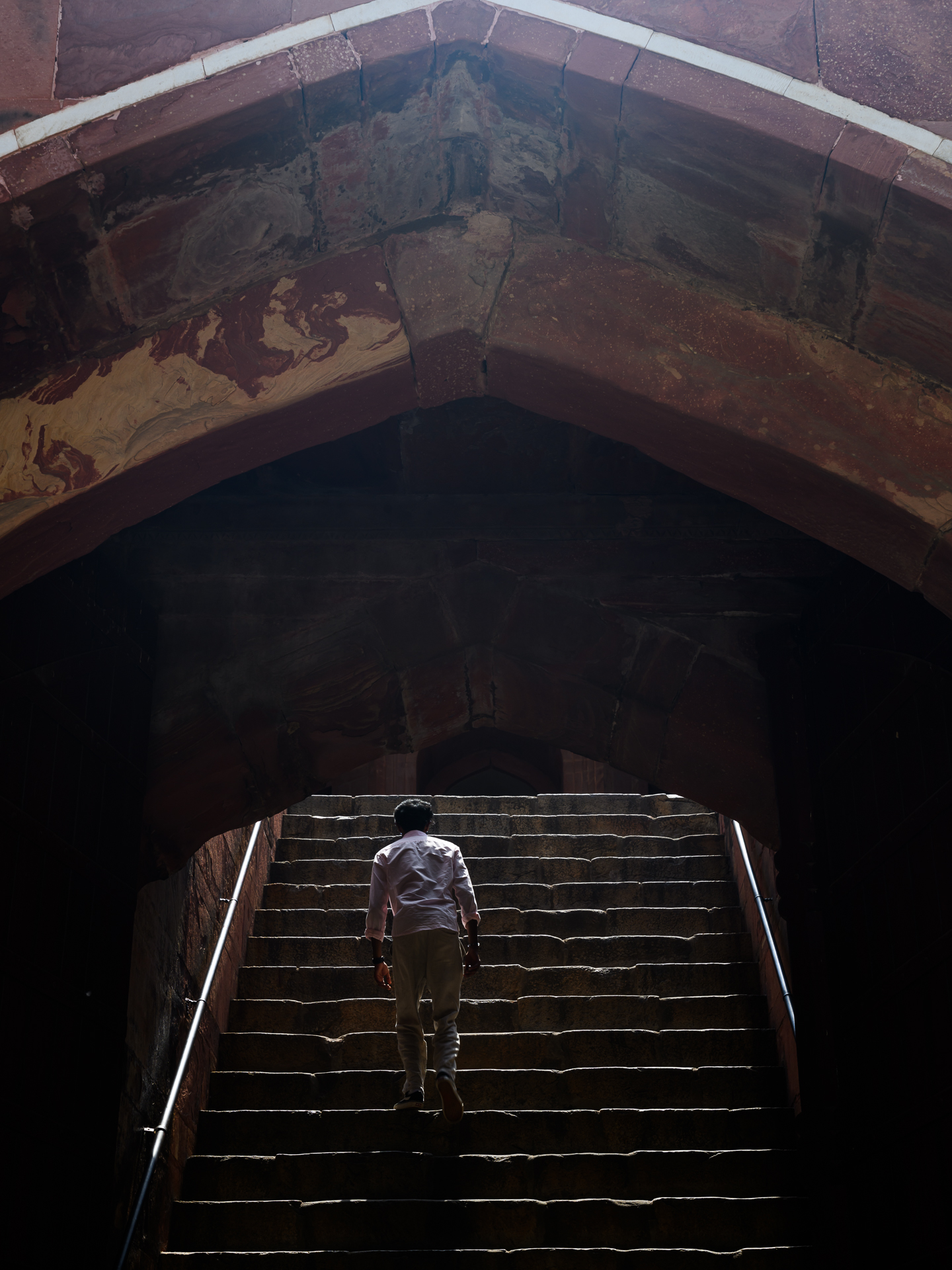 A man climbs stone steps from a dark arched interior towards a strip of bright sunlight.
