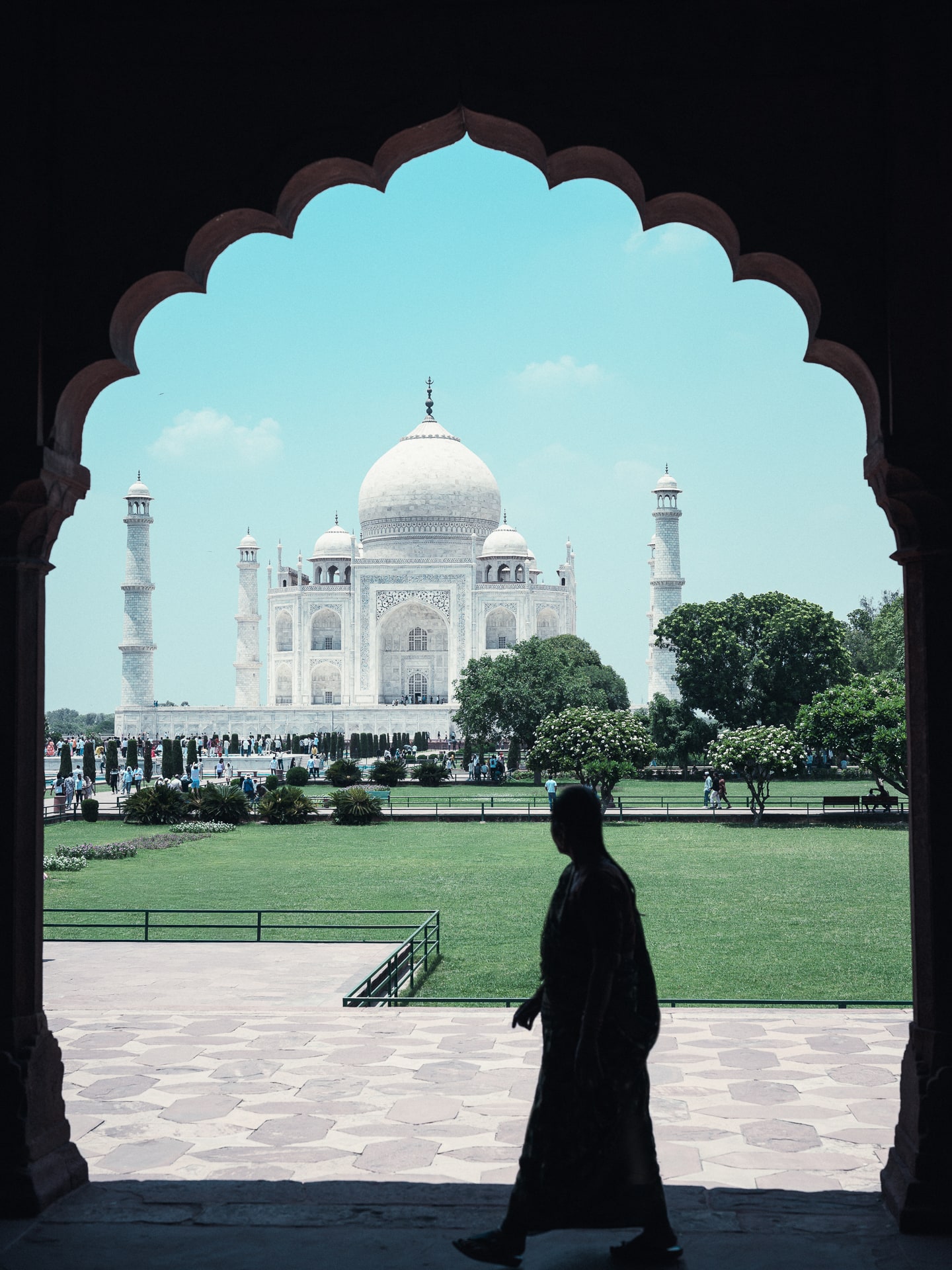 The Taj Mahal seen through a large cusped arch, with a person walking in silhouette across the marble foreground.