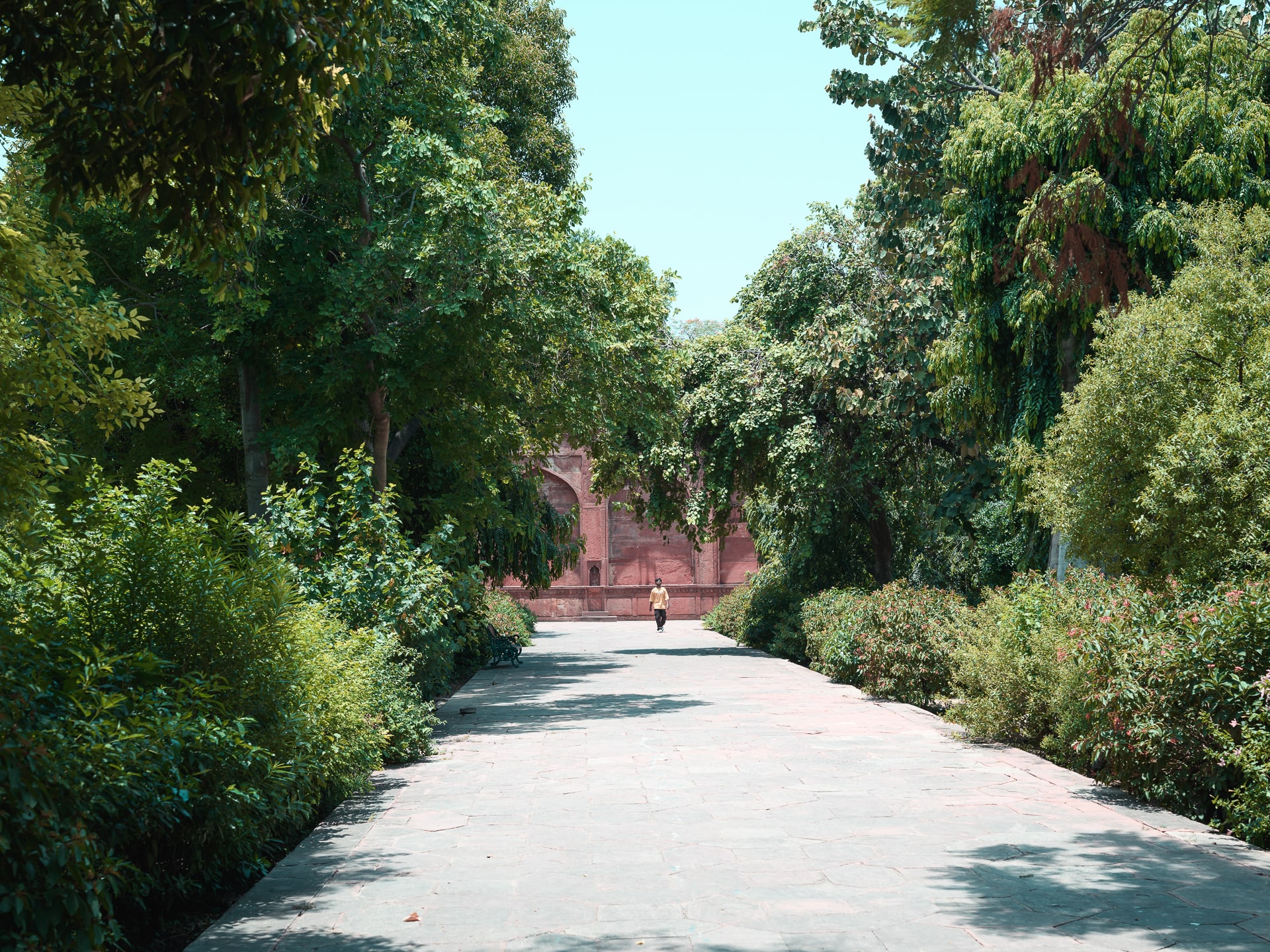 A tree-lined stone path leads towards a red sandstone gateway, with a lone figure in the distance.