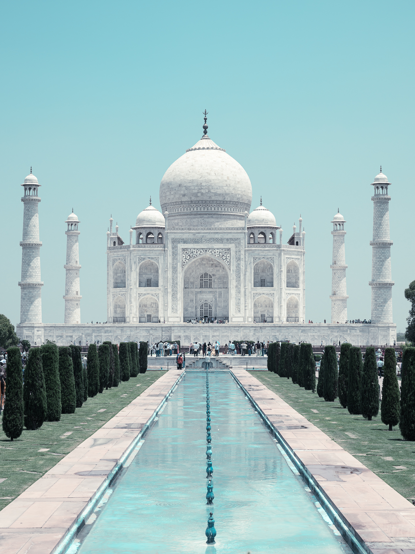 The Taj Mahal centred beyond its long reflecting pool beneath a pale blue sky.