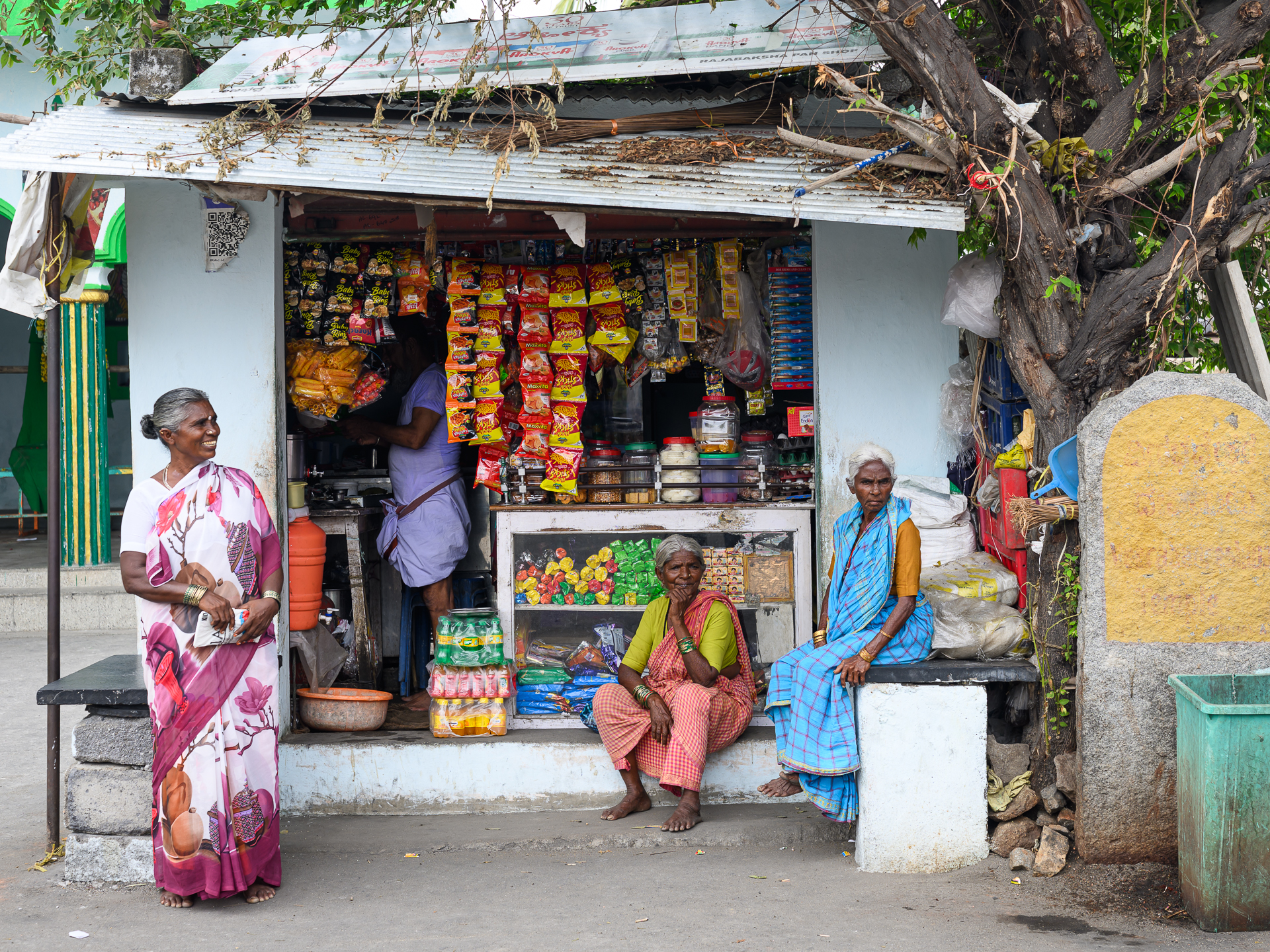 Four women gather around a small roadside shop filled with colourful hanging packets.