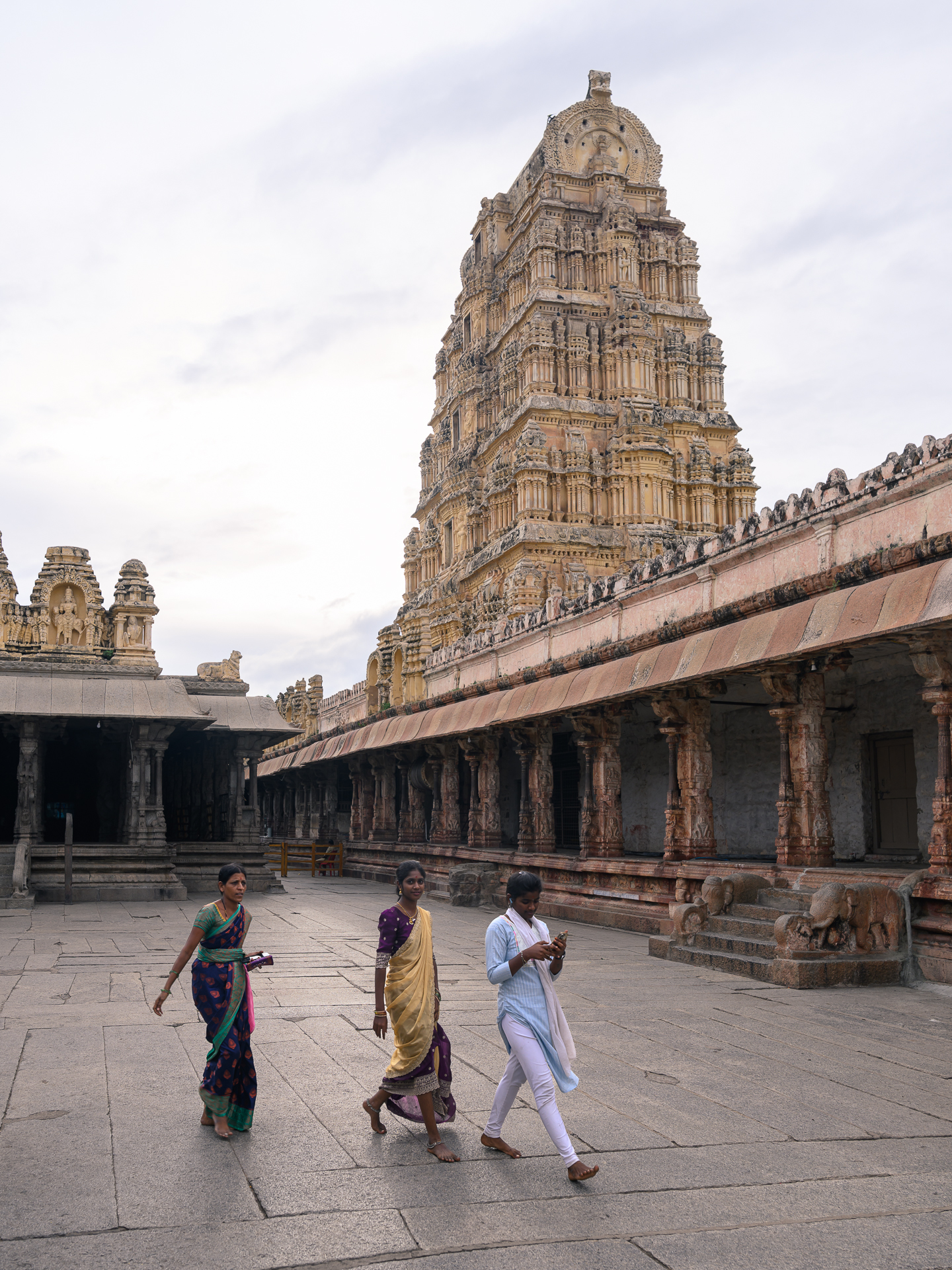 Three women cross a stone temple courtyard below a tall carved gopuram.