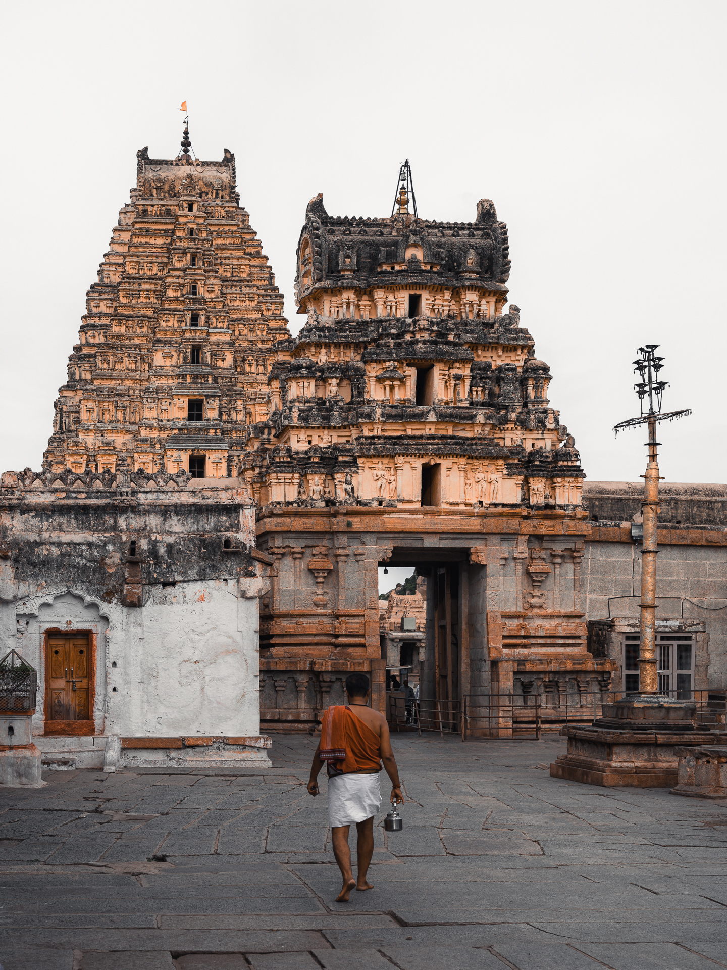 A barefoot man carrying a metal vessel walks towards a carved temple gateway.